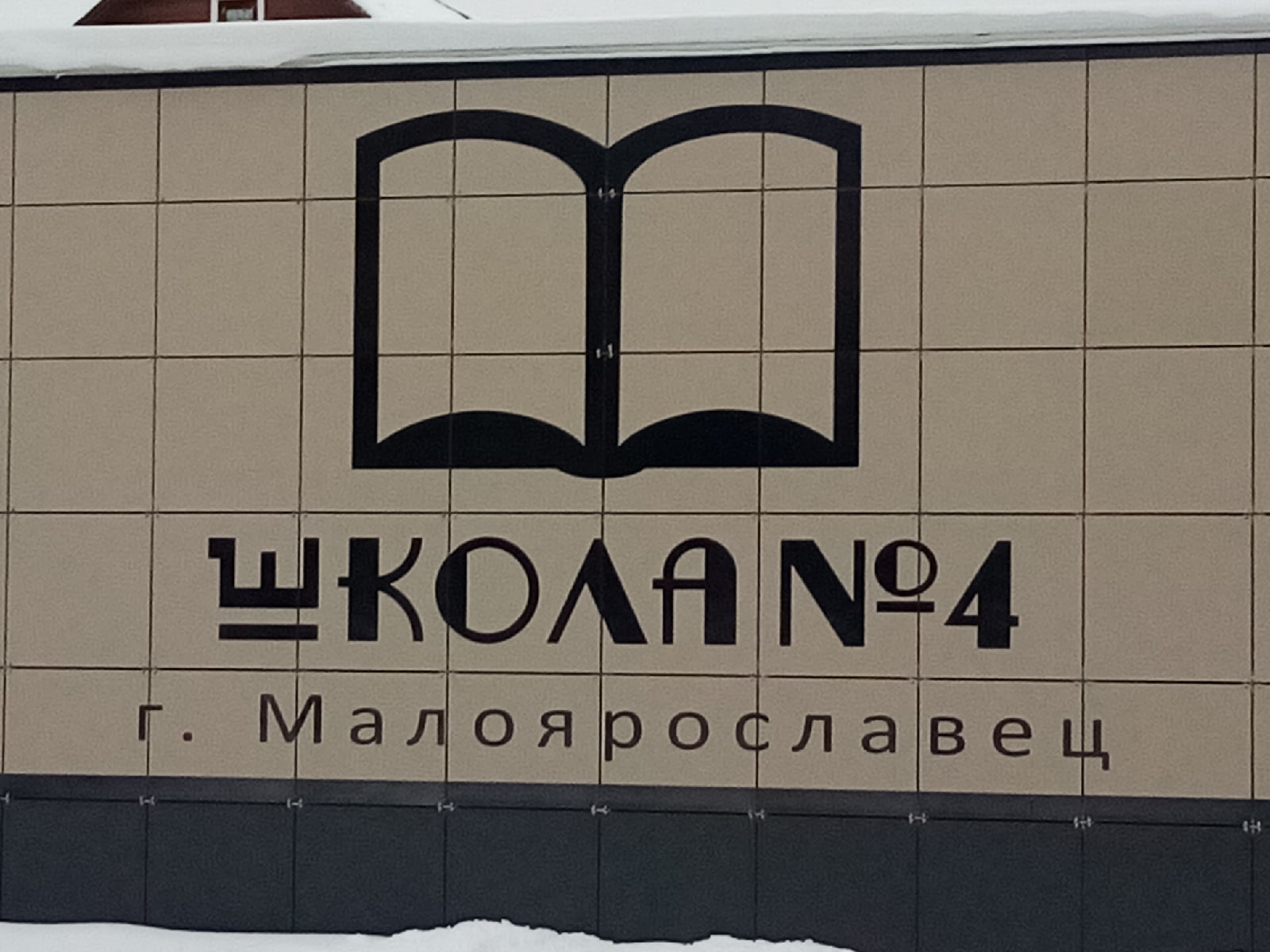 На стене здания школы изображена черно-белая эмблема. Табличка с надписью «Школа №4 г. Малоярославец». Вверху значок открытой книги. Фон состоит из плиток.